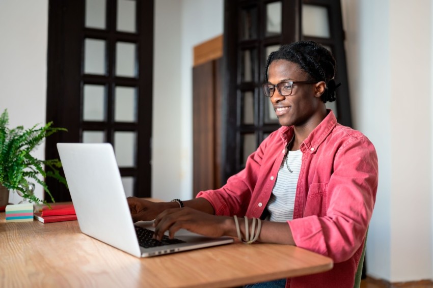 Happy satisfied young African guy freelancer sitting at home office enjoying working remotely